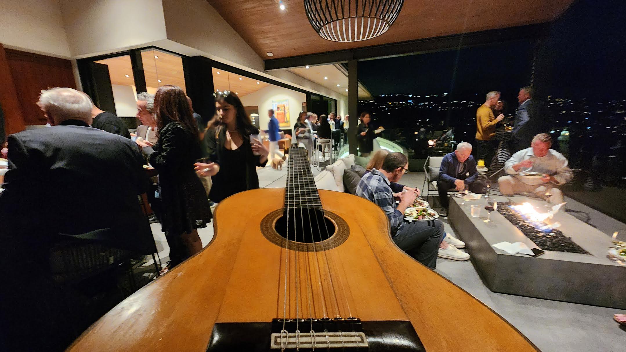 Guests in evening clothes gathered around a glowing fire pit, a nylon-string guitar in the near foreground